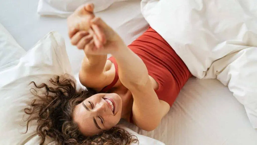 Top view of cheerful woman waking up after sleep and stretching on bed. High angle view of beautiful girl lying on bed and stretching after wake up. Awake woman wake up in morning and feeling fresh after a good rest.